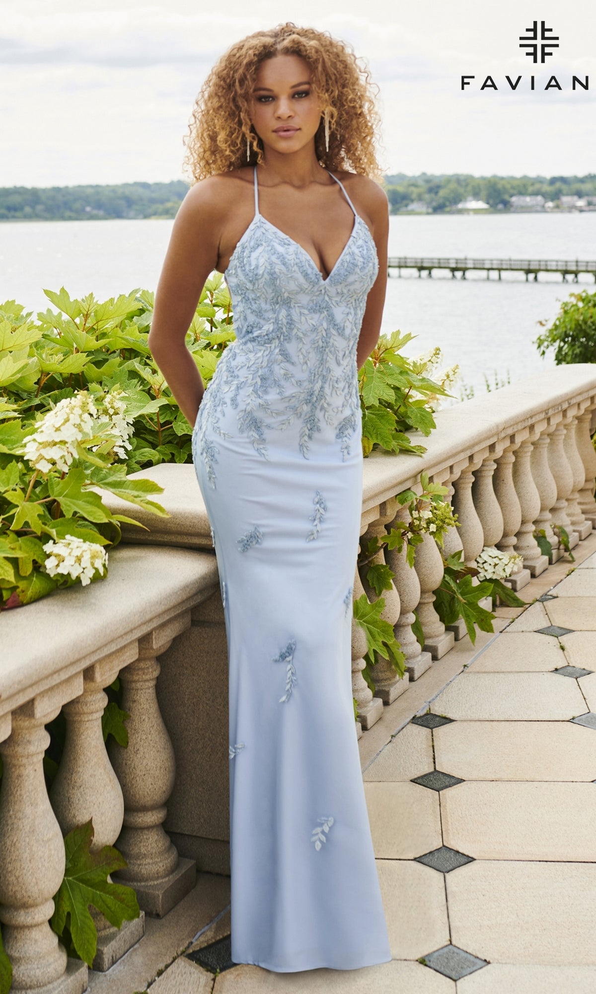 A woman in a fitted light blue Beaded Applique Long Faviana Prom Dress 11003 stands outdoors by a stone railing with water and greenery behind her. The Faviana logo is visible in the top right corner.